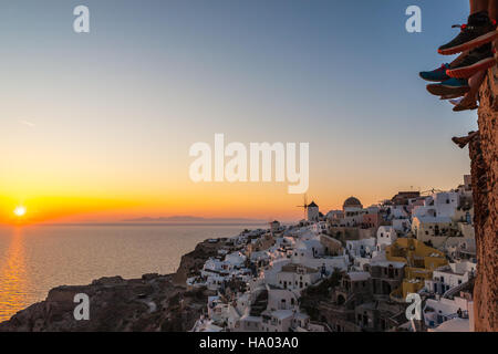 I turisti seduti su una parete con gambe penzolanti a guardare il tramonto di Oia - Santorini, Grecia Foto Stock