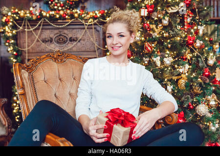 Giovane bella bionda donna seduta su una sedia vicino a albero di Natale e tenendo un dono. Nuovo anno. Foto Stock