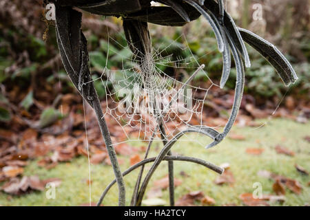 Autunnale coperto di brina ragnatela sulla statua di metallo con caduto foglie marroni sull'erba in background Foto Stock