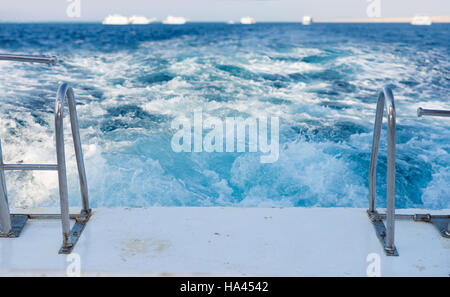 Vista di una nave della scia di onda tra metallo acciaio scale sul retro pontatura in teak di uno yacht a motore in un mare turchese Foto Stock