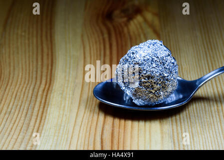 Sfera di eroina avvolti in un foglio di stagno sfera poggiante su un cucchiaino di argento su una tavola di legno superficie testurizzata. Foto Stock