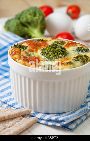 Dieta sana prima colazione. Broccoli casseruola con uova e formaggio in una ciotola bianco, pomodori freschi, pane fresco sul bianco sullo sfondo di legno Foto Stock