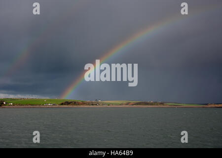 Brillante arcobaleno doppio sopra la tomaia cleddau in pembrokeshire contro un cielo scuro su acque calme Foto Stock