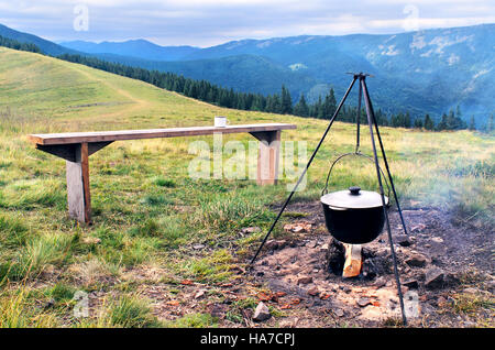 Camping fire e turisti bowler con la preparazione di cibo in montagna. La cottura a fuoco in condizioni di campo. Falò con bowler cena. La cottura in marzo Foto Stock
