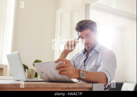 Colpo di un giovane uomo seduto a tavola alla ricerca di documenti e di pensare. Uomo d'affari andando attraverso documenti all ufficio di casa. Foto Stock