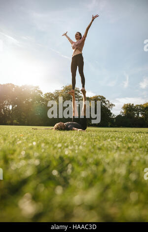 Colpo verticale di adattamento coppia giovane facendo esercizio acroyoga nel parco. Uomo disteso su erba e donna di bilanciamento. Foto Stock