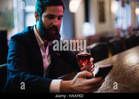 Imprenditore utilizzando il telefono cellulare con un bicchiere di vino rosso in mano Foto Stock