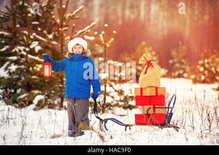 Carino piccolo ragazzo in Santa hat holding lanterna di Natale e porta una slitta di legno pieno di confezioni regalo in boschi innevati. Xmas, neve e divertimento invernale per fam Foto Stock