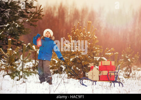 Carino piccolo ragazzo in Santa hat holding lanterna di Natale e porta una slitta di legno pieno di confezioni regalo in boschi innevati. Xmas, neve e divertimento invernale per fam Foto Stock