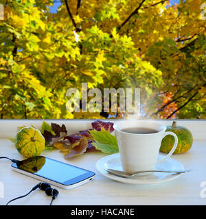 Tazza di caffè, lo smartphone, le cuffie e le foglie di autunno su un davanzale. Nella finestra di sfondo con paesaggio autunnale Foto Stock
