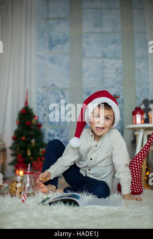 Carino per bambini in età prescolare, ragazzo, la lettura di un libro e mangiare i biscotti a casa mentre nevicava all'aperto Foto Stock