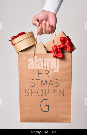 Primo piano della mano di un giovane uomo caucasico tenendo una carta shopping bag pieno di doni e il testo lo shopping di Natale scritta in esso la formazione di un Cristo Foto Stock