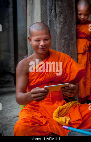 Sorridente monaco buddista nel tradizionale tunica arancione guardando il suo smartphone in Angkor Wat a Siem Reap, Cambogia con piccolo ragazzo in background. Foto Stock