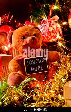 Primo piano di un orso di peluche con una lavagna con il testo joyeux noel, buon natale scritto in francese e alcuni doni sotto un albero di natale ornamento Foto Stock