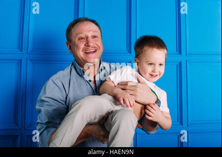 Ridendo nonno con il nipote come giocano insieme al chiuso nella stanza vivente con la cute giovane ragazzo che abbraccia Lui da dietro Foto Stock