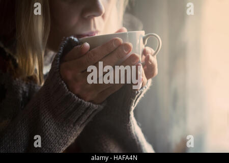 La donna in grigio felpa lavorata a maglia con motivi tradizionali bevendo una tazza bianca di caffè caldo con vapore che fuoriesce nella luce del mattino Foto Stock