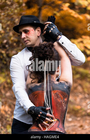 Giovane uomo ad imitazione delle prestazioni musicali sulla donna torna con il dipinto di violoncello. Colorato parco di alberi sullo sfondo Foto Stock