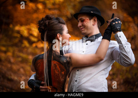 Giovane uomo ad imitazione delle prestazioni musicali sulla donna torna con il dipinto di violoncello. Colorato parco di alberi sullo sfondo Foto Stock