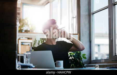 Colpo di giovane uomo seduto al suo ufficio parlando al cellulare. Business man lavorando alla sua scrivania con computer portatile effettuando una chiamata telefonica. Foto Stock