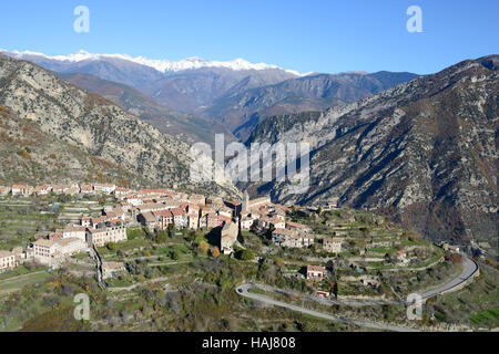 VISTA AEREA. Villaggio in cima alla collina che domina la valle Vésubie con le Alpi del Mercantour innevate in lontananza. Utelle, Alpi Marittime, Francia. Foto Stock