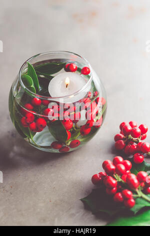 Natale Decorazioni per la tavola. Vaso di vetro con agrifoglio bacche e bianco candela galleggiante. Foto Stock