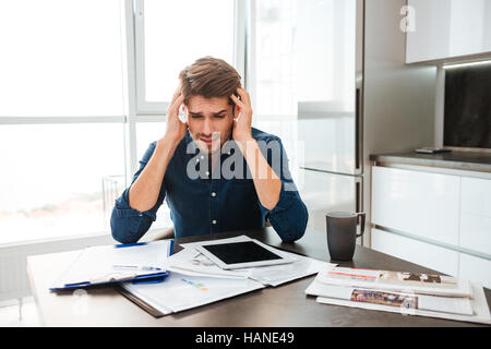 Giovane uomo confuso analizzando le finanze tenendo la testa con le mani. Seduto vicino a tavola con tablet e documenti. Foto Stock