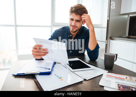 Giovane uomo confuso analizzando le finanze a casa mentre si tiene la testa con la mano e guardando i documenti. Seduto vicino a tavola con tablet. Foto Stock