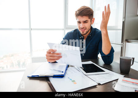 Giovane uomo triste analizzando le finanze di casa mentre gesticolando con la mano e guardando i documenti. Seduto vicino a tavola con tablet. Foto Stock