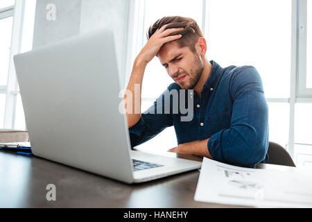 Foto di confuse giovane uomo seduto vicino a laptop e tenendo la testa con la sua mano. Gli occhi chiusi. Foto Stock