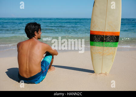 Vista posteriore del tiro del giovane uomo seduto sulla spiaggia con la tavola da surf. Surfista maschio rilassante in riva al mare su un giorno d'estate. Foto Stock