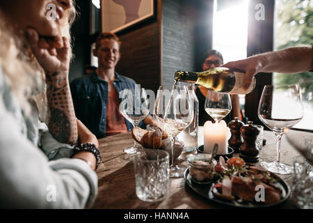 Man mano versando il vino bianco dalla bottiglia in bicchieri con gli amici seduti attorno al tavolo. Un gruppo di giovani con cibo e bevande a restau Foto Stock