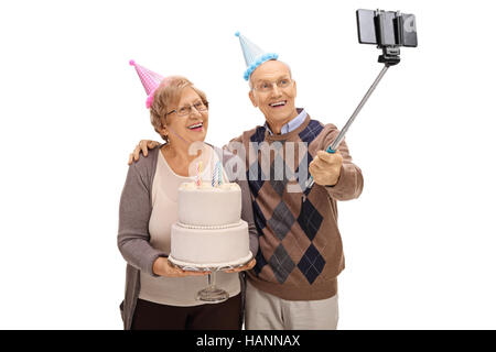 Seniors gioiosa festa con cappelli e una torta di compleanno prendendo un selfie con un bastone isolato su sfondo bianco Foto Stock