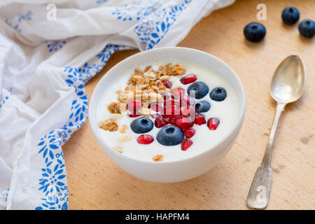 Lo yogurt naturale con granola o muesli, mirtilli freschi e i semi di melograno in bianco ciotola sul tavolo di legno Foto Stock