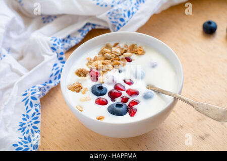 Lo yogurt naturale con granola o muesli, mirtilli freschi e i semi di melograno in bianco ciotola sul tavolo di legno Foto Stock