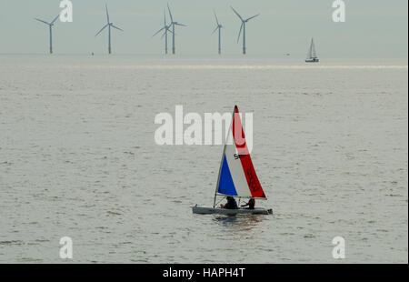 Una piccola barca a vela dinghy contenente i giovani sul mare, con turbine eoliche visibile in background. Foto Stock