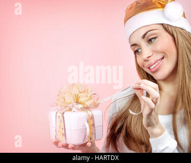 Ritratto di una donna allegra indossando golden Santa hat tenendo le mani in confezione regalo e la sua apertura, Cute girl su sfondo rosa, foto con spazio di copia Foto Stock