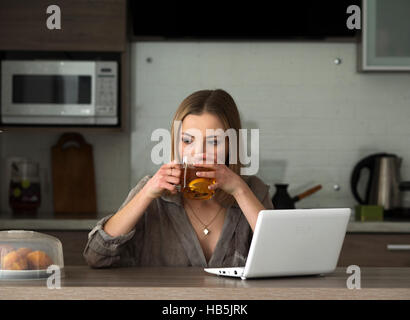 Giovane bella donna caucasica drinkig tè e cerca informazioni in social media su net-book Foto Stock