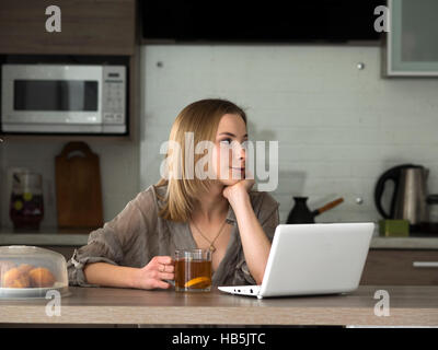 Giovane bella donna caucasica drinkig tè e cerca informazioni in social media su net-book Foto Stock