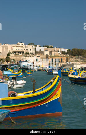 Colorate barche da pesca locali di Marsaxlokk sull'isola di Malta con uno sfondo di edifici lato porto e un cielo blu Foto Stock