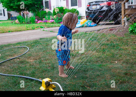 Caucasian Baby Boy gioca con un soffione di erogazione dell'acqua nel suo prato anteriore durante la stagione estiva Foto Stock