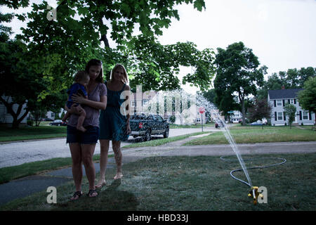 Caucasian Baby Boy tenuto da sua madre e zia dopo aver giocato con un soffione di erogazione dell'acqua nel suo prato anteriore durante la stagione estiva Foto Stock