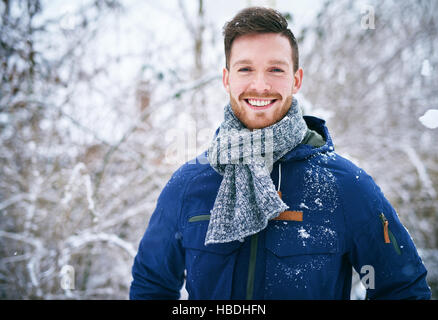 Giovane uomo barbuto in caldo cappotto sorridente alla fotocamera su sfondo sfocato dei boschi innevati. Foto Stock