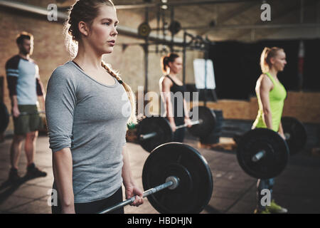 Muscolari gravi giovane donna con coda di cavallo e camicia grigia eseguendo sollevamento morto barbell esercizi con altri tre partecipanti Foto Stock