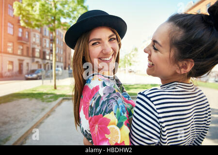 Moda giovane donna con un bel sorriso indossando un elegante piccolo hat in piedi insieme con un amico svolta a guardare la fotocamera in un contesto urbano Foto Stock