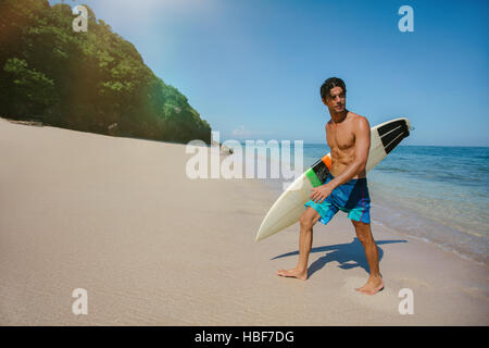 Colpo di giovane maschio surfer tenendo tavole da surf a piedi al di fuori del mare. Maschio caucasico con la tavola da surf a piedi lungo la spiaggia e guardando lontano. Foto Stock