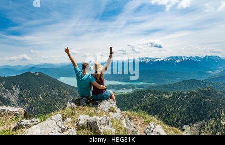 Coppia giovane con le braccia in aria, la vista del lago di Kochel dal picco Heimgarten, sinistra Herzogstand, Alta Baviera, Baviera, Germania Foto Stock