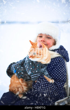 Bambina tiene un gatto rosso nelle sue mani seduto su una slitta in inverno. Foto Stock