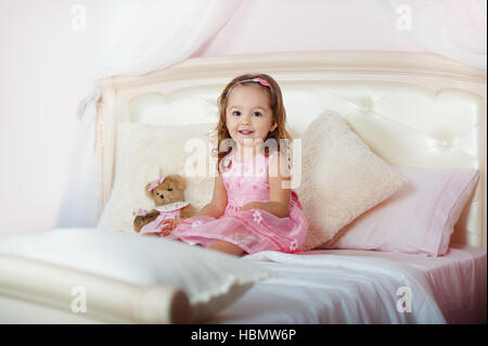 Bella bimba bionda in abito rosa con Teddy bear seduta su di un letto per bambino e ride rumorosamente nel luminoso interno della casa Foto Stock