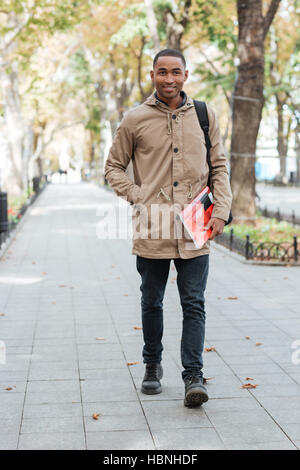 Immagine dell Africa uomo gioioso indossando uno zaino di camminare sulla strada tenendo libro e guardando la fotocamera. Foto Stock