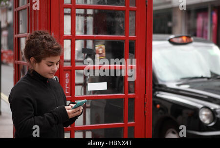 16 enne adolescente sorrisi come lei utilizza uno smartphone e sorrisi al messaggio di testo come ella si appoggia contro un vuoto di Londra cabina telefonica Foto Stock